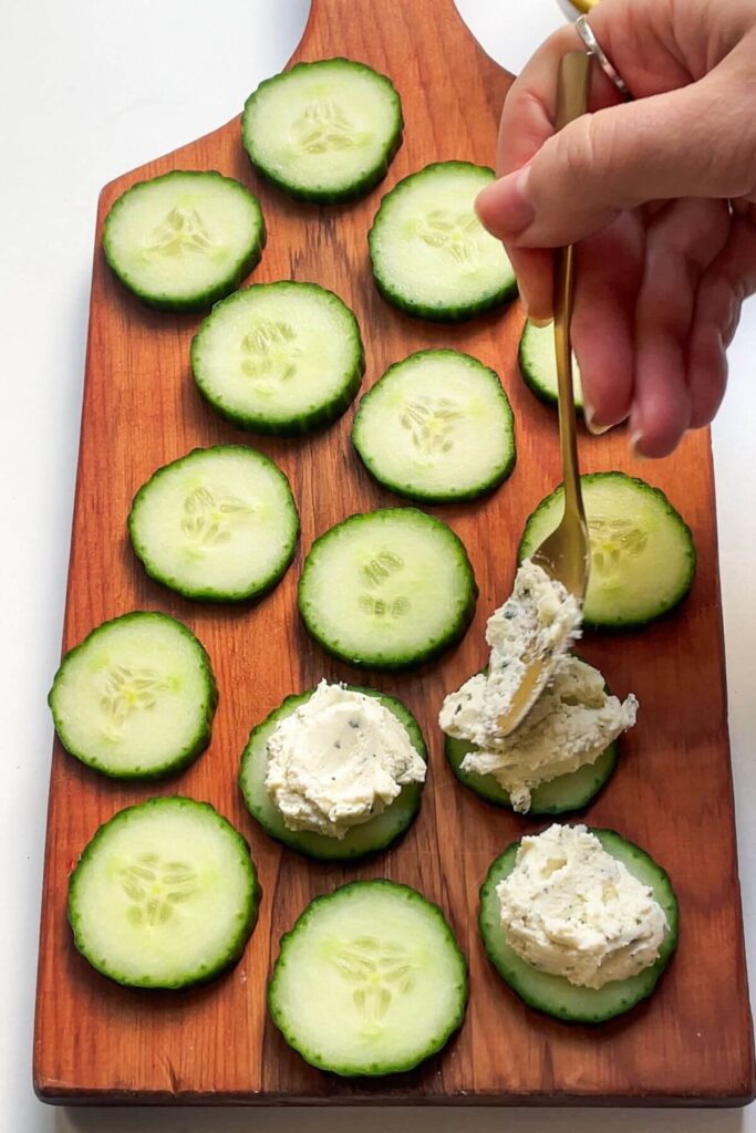 Spooning whipped boursin onto cucumber slices on a wooden board.