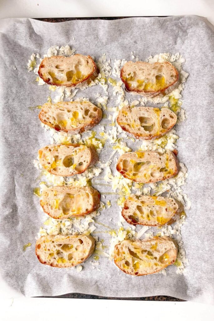 Baguette slices on top of grated cheese on a lined baking tray.