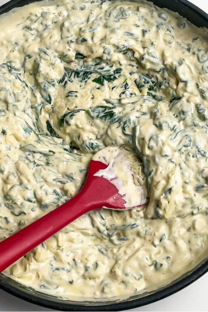 Red spatula stirring grated cheese into creamy spinach dip in a pan.