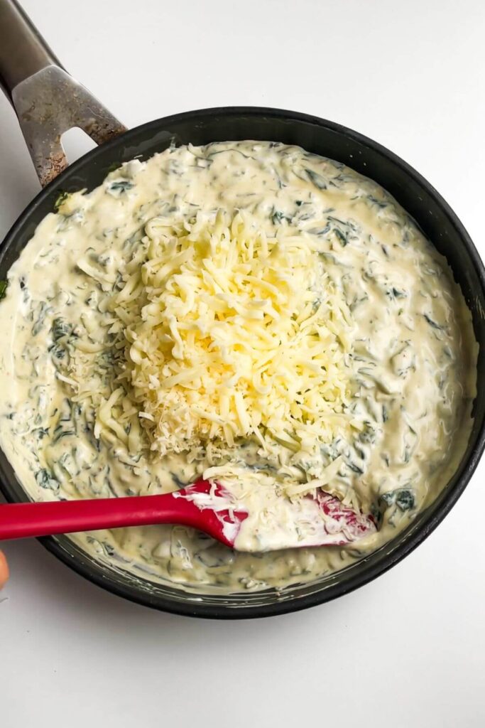 Grated cheese added to creamy spinach dip in a small pan.