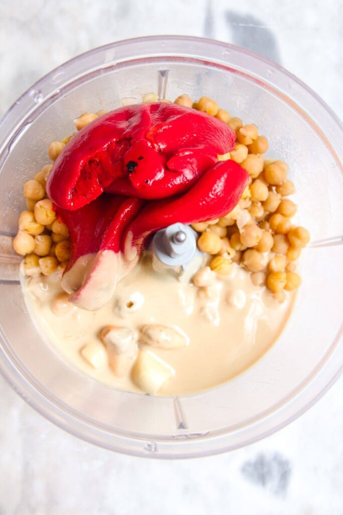 Chickpeas, tahini and red pepper in the bowl of a food processor.