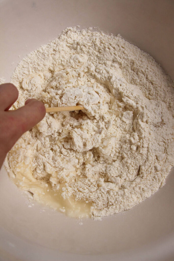 Hand holding a chopstick, mixing flour and water into a dough in a mixing bowl.