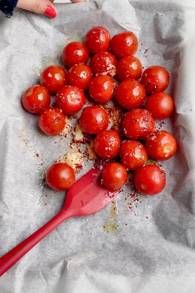 Red spatula mixing cherry tomatoes with garlic and olive oil on a lined tray.