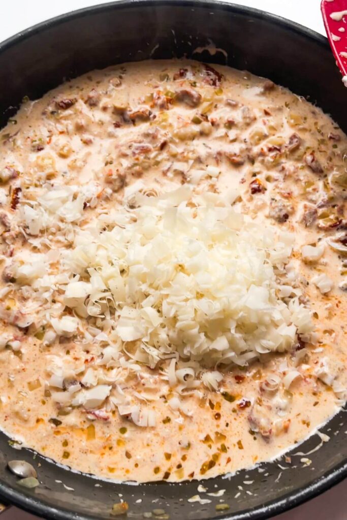 Grated parmesan added to sundried tomatoes in a creamy sauce in small pan.