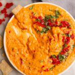 Cracker spread through a small white plate with red pepper hummus on it.