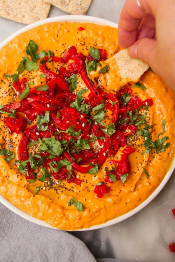 Hand scooping cracker in red pepper hummus on a white plate.