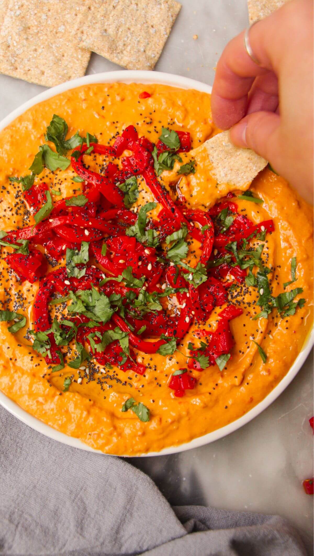 Hand scooping cracker in red pepper hummus on a white plate.