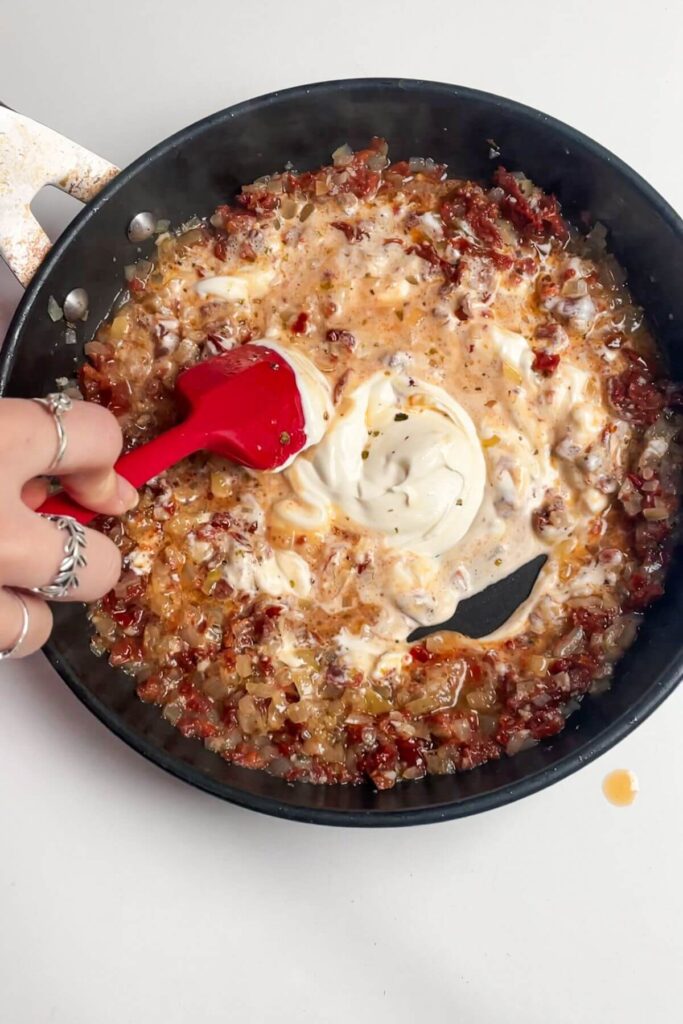 Red spatula stirring sour cream into sundried tomatoes in a small pan.