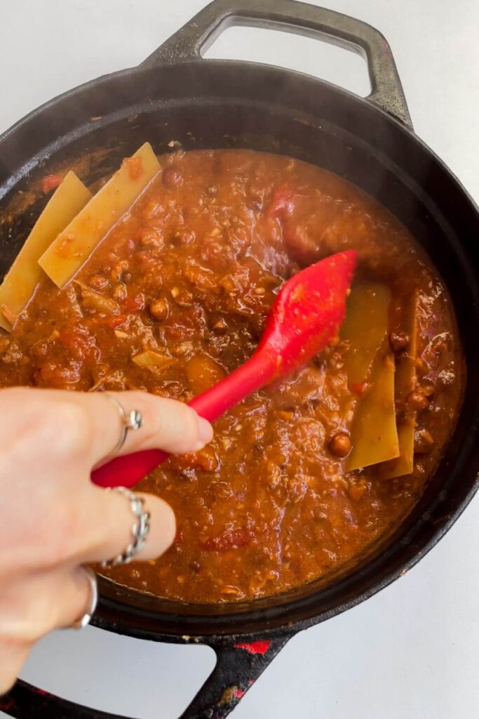 Hand holding spatula, stirring lasagna sheets through soup in a large pot.