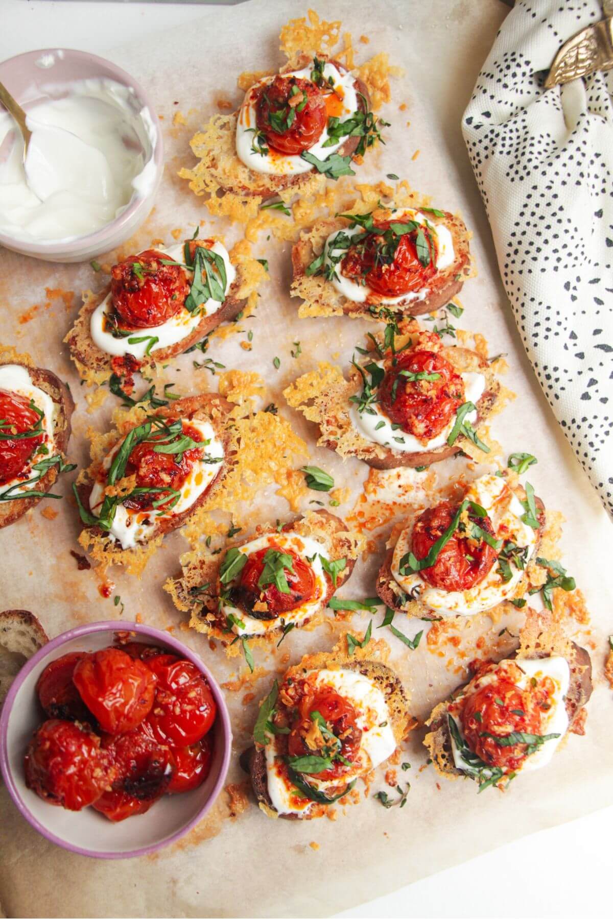 Cheesy crusted roasted tomato crostini on a wooden borad with more tomatoes on the side.