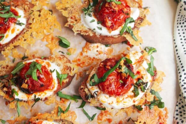 Cheesy crusted roasted tomato crostini on a wooden borad with more tomatoes on the side.