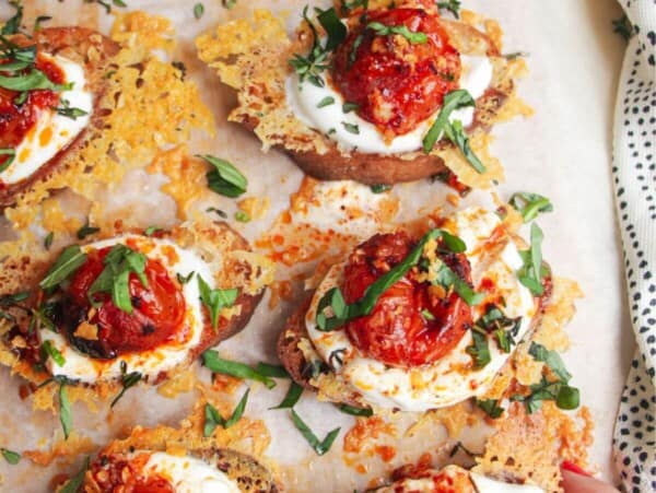 Cheesy crusted roasted tomato crostini on a wooden borad with more tomatoes on the side.