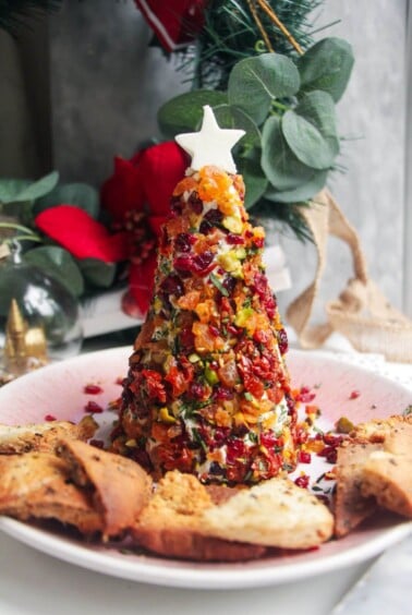Coated Christmas tree cheese ball topped with a cheese star on a pink plate with pita chips on the side.