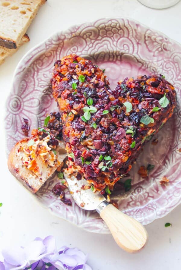 Heart shaped cheese ball, covered in dried fruit on a small pink plate, with bread on the side.