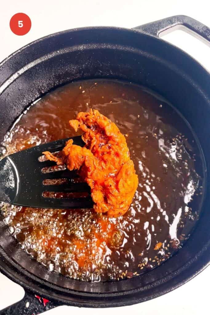 Spatula holding up fried chicken tender with pot of chicken frying behind.
