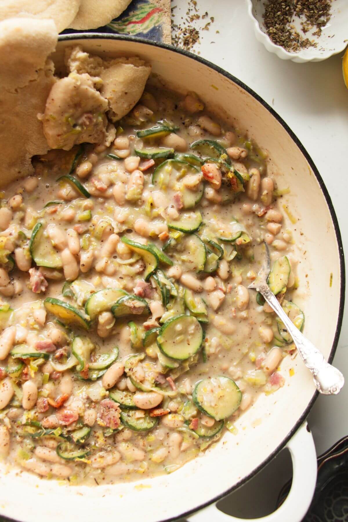 Creamy leek zucchini beans in a large pot with a silver spoon and pita bread on the side.