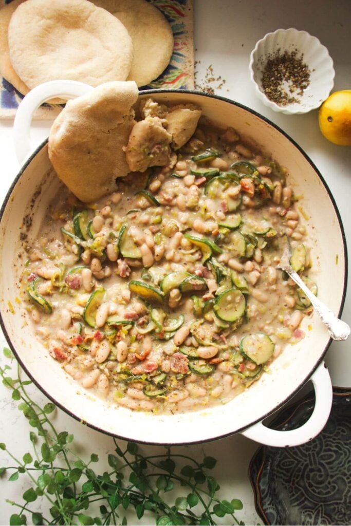 Creamy leek zucchini beans in a large pot with a silver spoon and pita bread on the side.