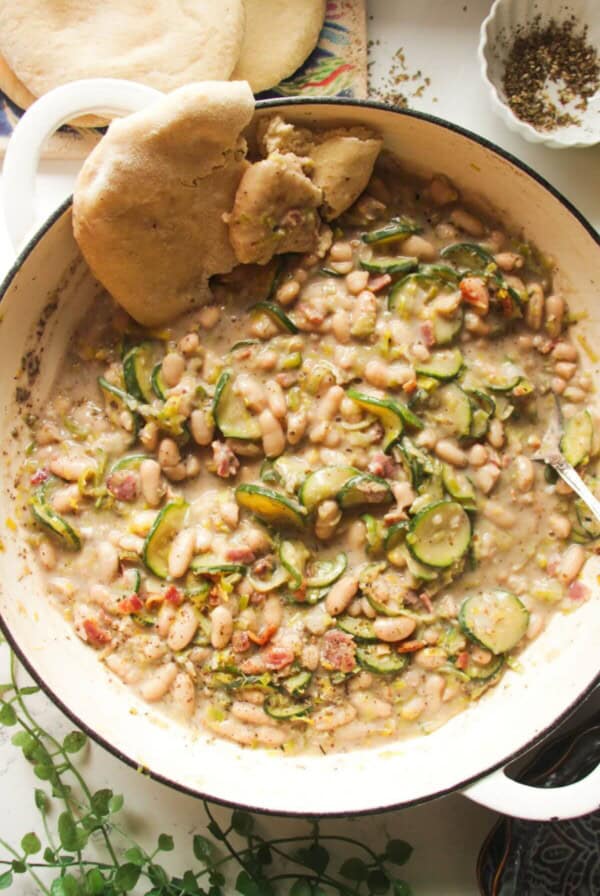 Creamy leek zucchini beans in a large pot with a silver spoon and pita bread on the side.