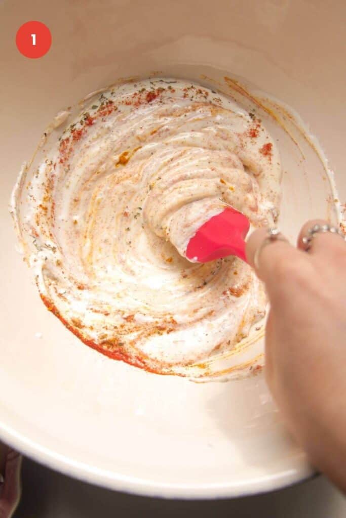 Hand holding spatula, mixing chicken marinade in bowl.