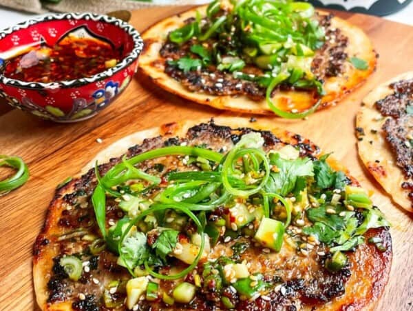 Three dumpling tacos on a wooden board with chilli oil on the side.
