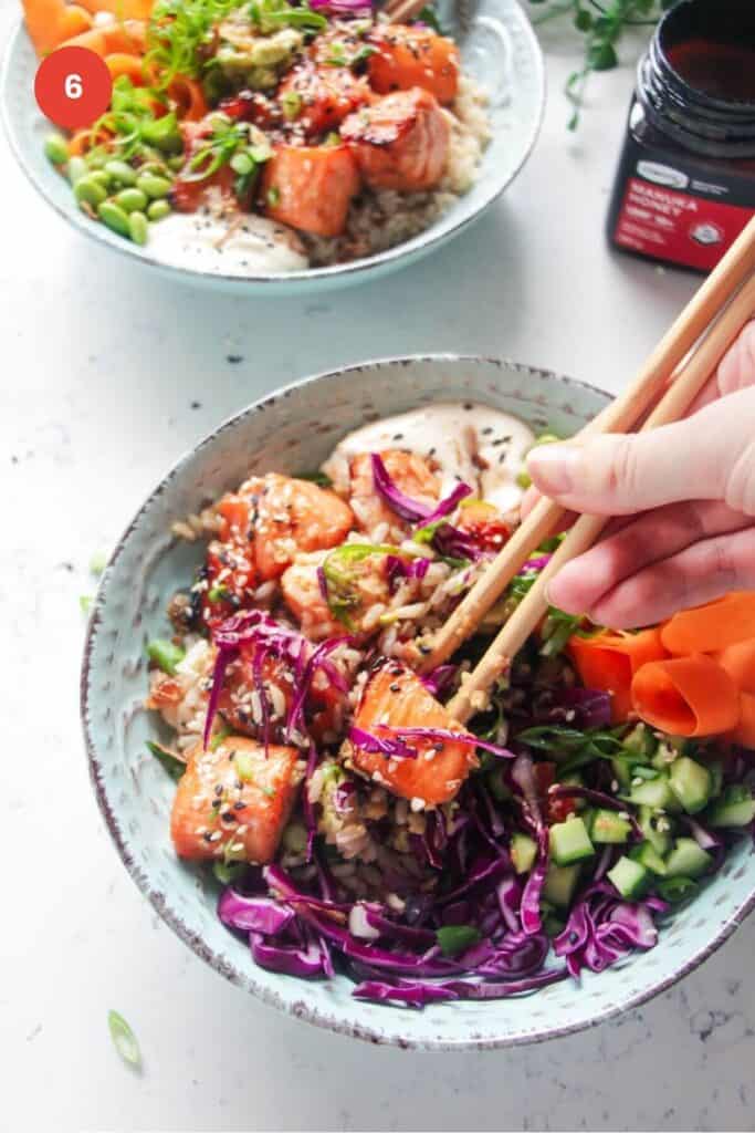 Hand holding chopsticks picking up salmon cube from poke bowl.
