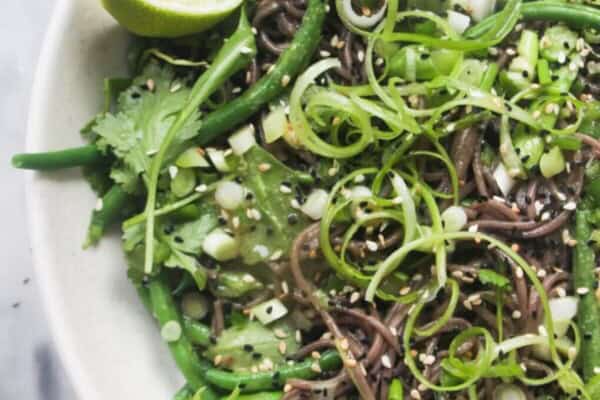 Cold Asian soba noodle salad with lots of green vegetables in a large serving platter, with jug of dressing on the side.