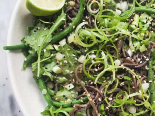 Cold Asian soba noodle salad with lots of green vegetables in a large serving platter, with jug of dressing on the side.