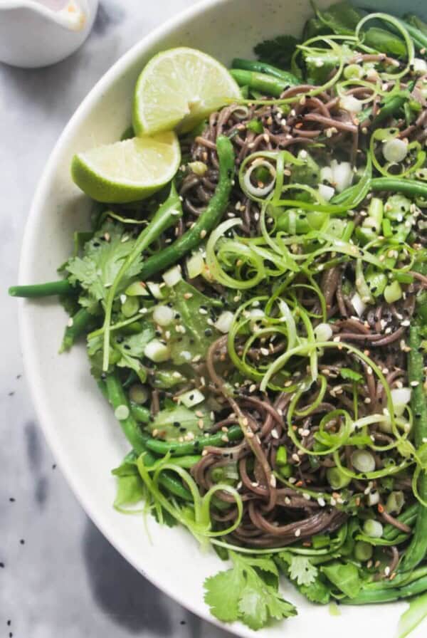 Cold Asian soba noodle salad with lots of green vegetables in a large serving platter, with jug of dressing on the side.