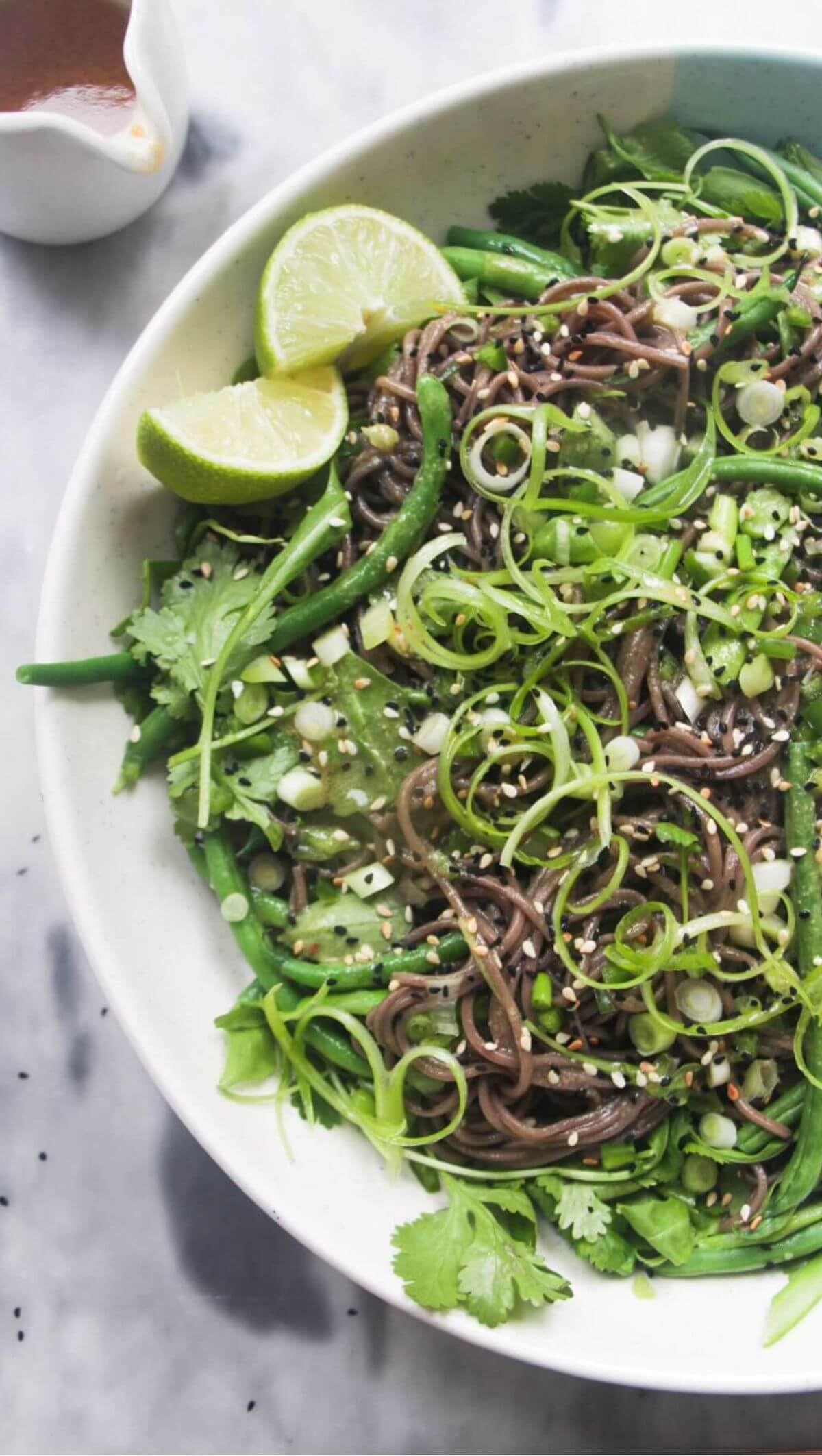 Cold Asian soba noodle salad with lots of green vegetables in a large serving platter, with jug of dressing on the side.