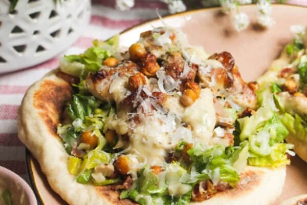 Flatbread on a plate topped with Caesar salad, charred chicken pieces and creamy Caesar dressing.