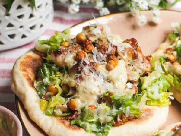 Flatbread on a plate topped with Caesar salad, charred chicken pieces and creamy Caesar dressing.