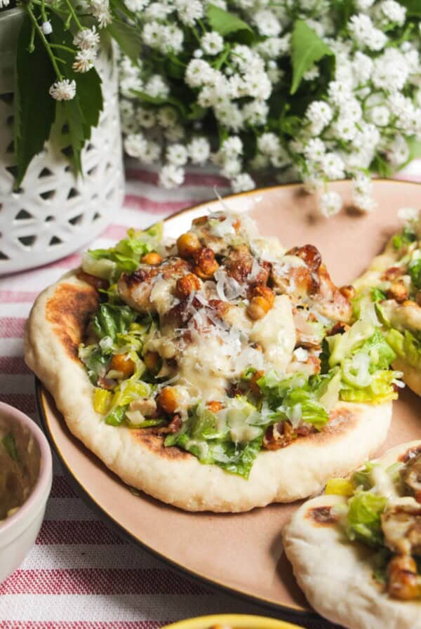 Flatbread on a plate topped with Caesar salad, charred chicken pieces and creamy Caesar dressing.