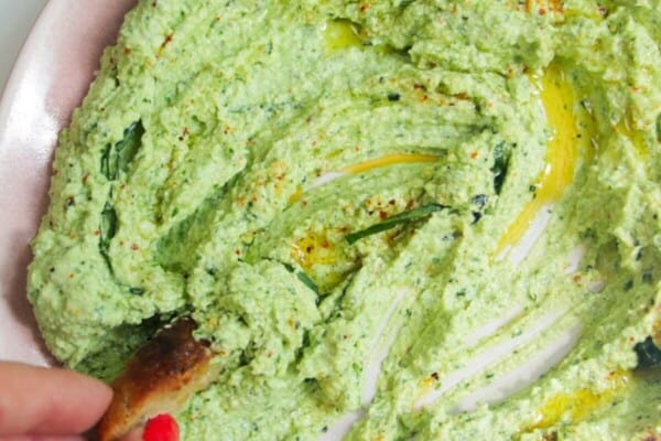 Whipped feta pesto dip on a pink plate with a small piece of bread on thes side.