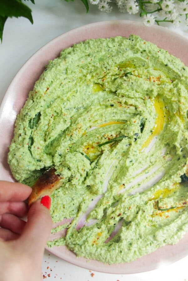 Whipped feta pesto dip on a pink plate with a small piece of bread on thes side.