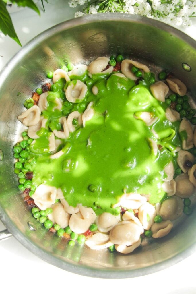 Spinach basil sauce added to cooked pasta, peas and pancetta in a pan.