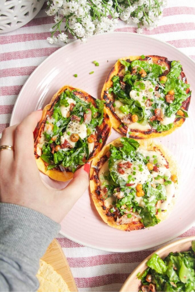 Three chicken Caesar tacos on a pink plate with a hand reaching out to take one.