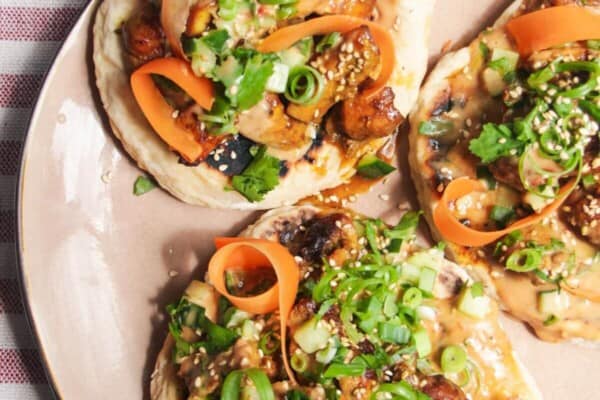 Three chicken satay flatbreads on a pink plate, topped with herbs and peanut sauce.