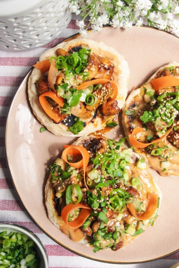 Three chicken satay flatbreads on a pink plate, topped with herbs and peanut sauce.