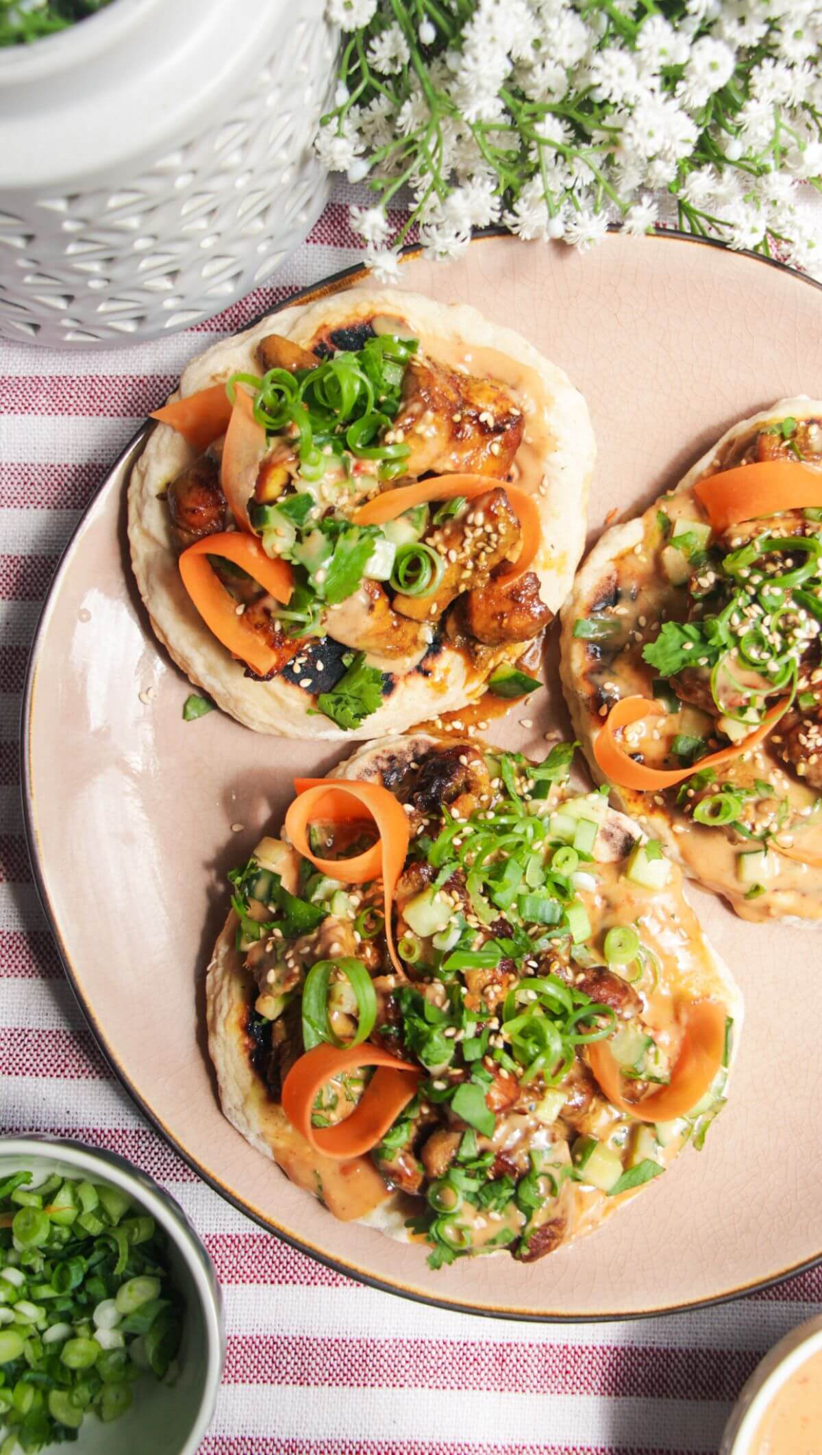 Three chicken satay flatbreads on a pink plate, topped with herbs and peanut sauce.