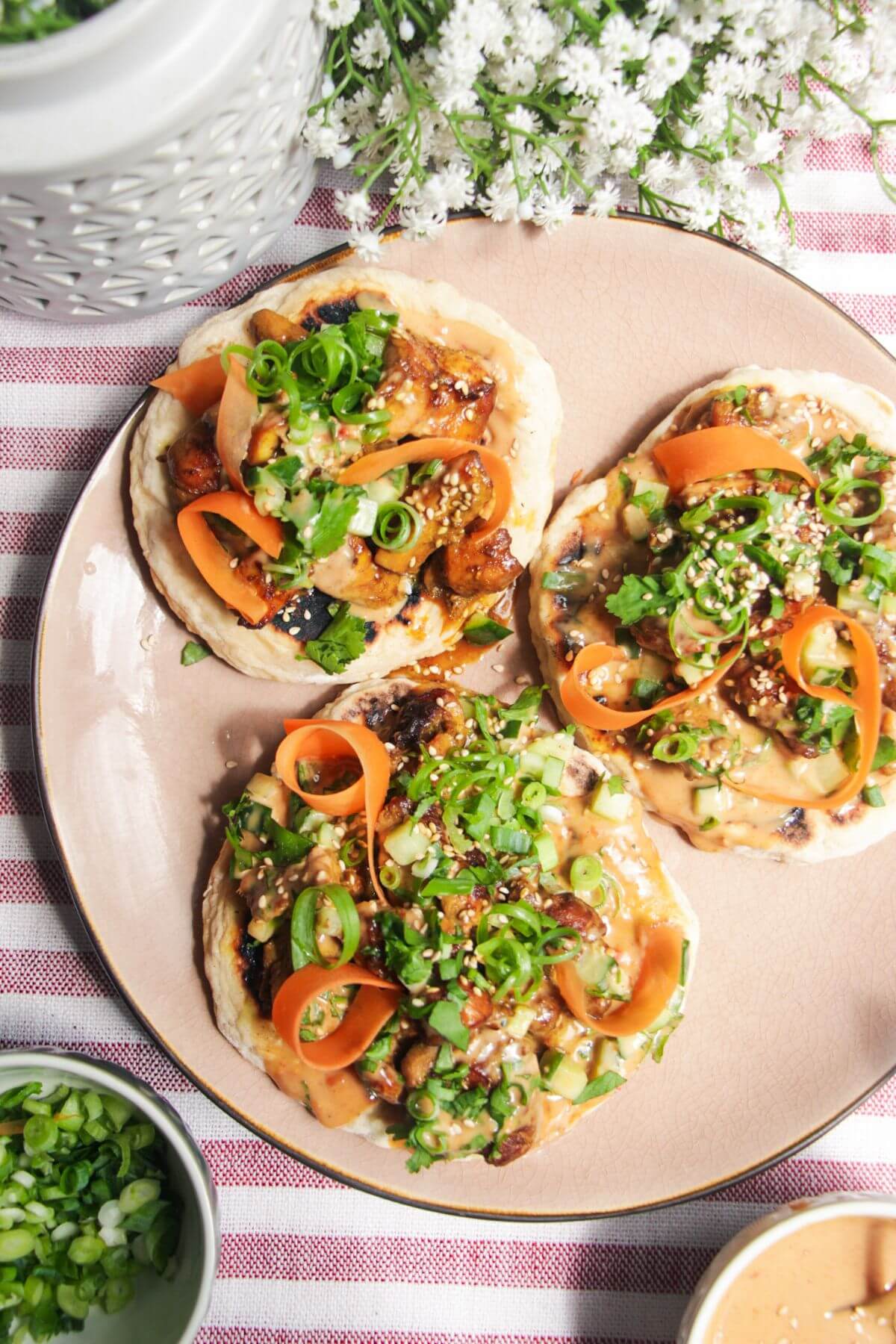 Three chicken satay flatbreads on a pink plate, topped with herbs and peanut sauce.