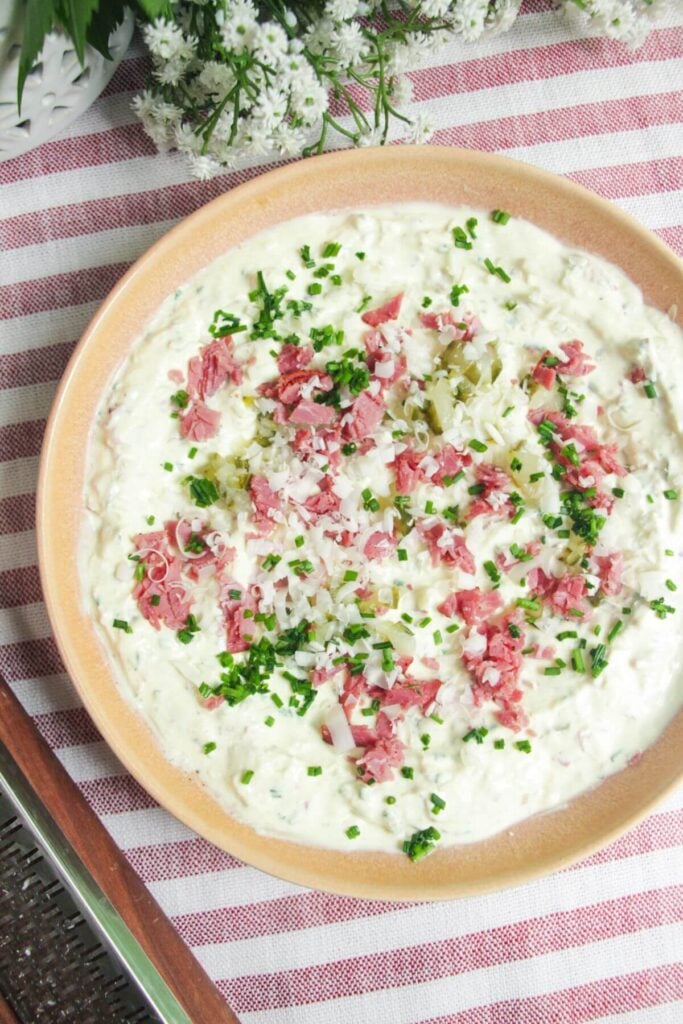 Creamy pastrmi dip in a pink bowl, topped with pickles, pastrami and gruyere cheese.