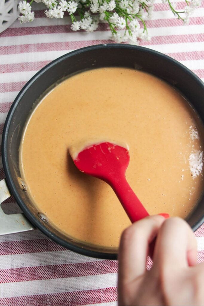 Hand holding red spatula stirring peanut sauce in a pan.