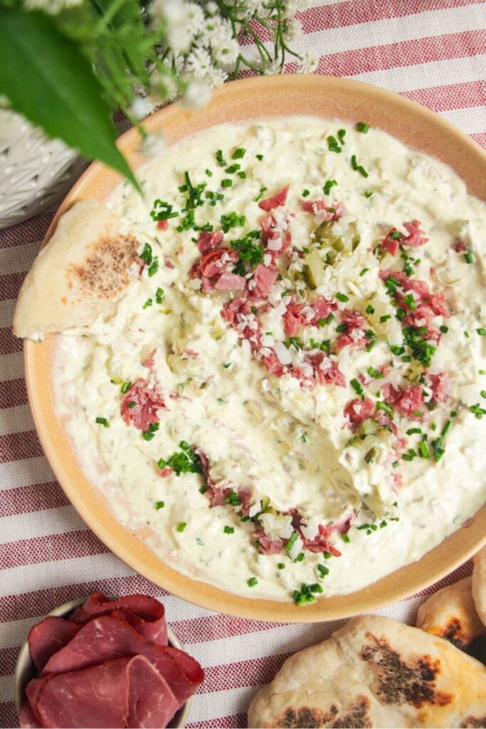 Creamy pickle dip in a pink bowl, topped with pickles, pastrami and gruyere cheese.