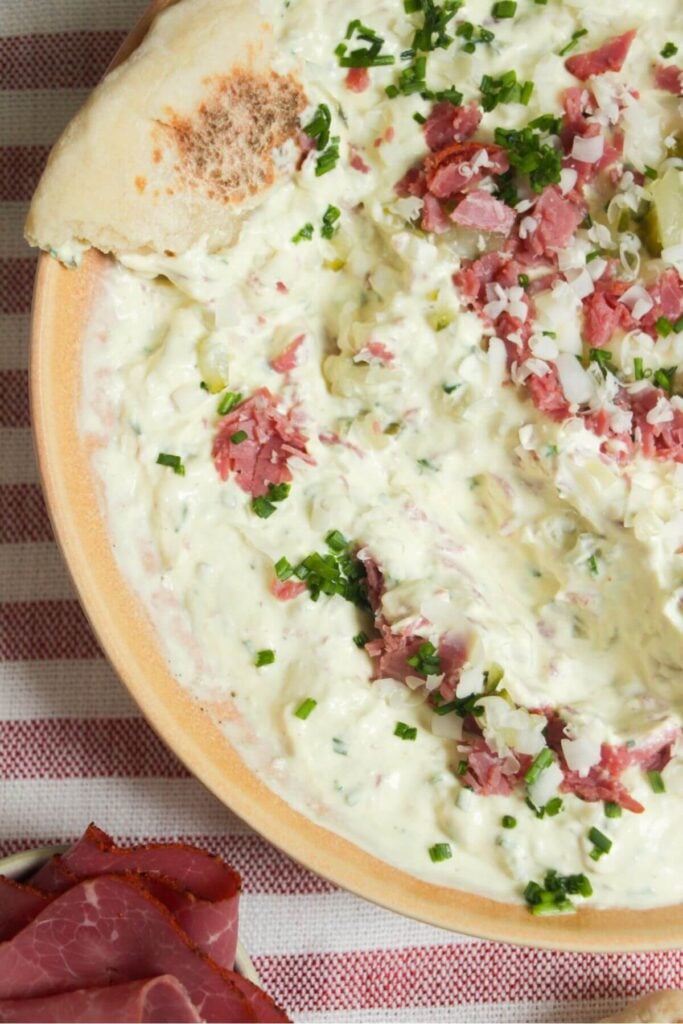 Creamy pickle dip in a pink bowl, topped with pickles, pastrami and gruyere cheese.