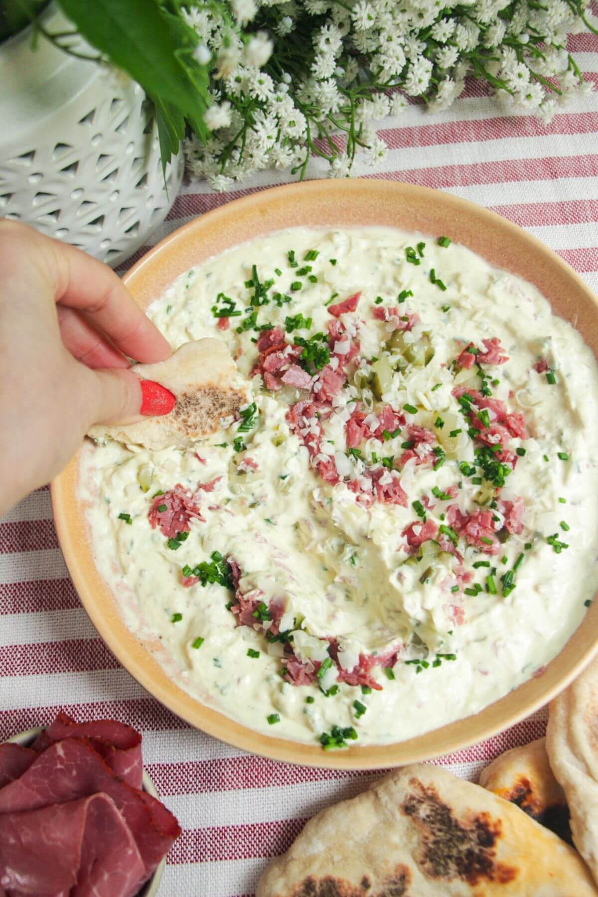 Creamy pickle dip in a pink bowl, topped with pickles, pastrami and gruyere cheese.
