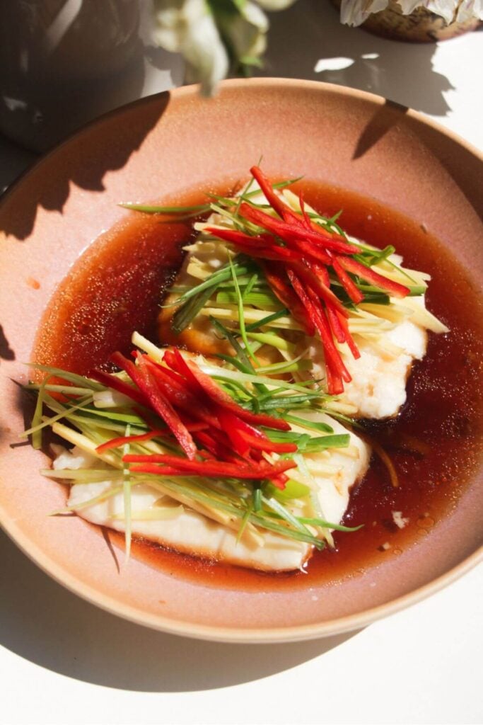 Soy sauce poured over fish fillets in pink bowl with scallions, chili and ginger batons on top.