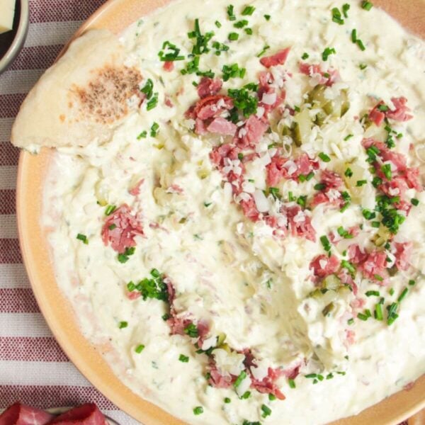 Creamy pickle dip in a pink bowl, with a piece of bread on the side.