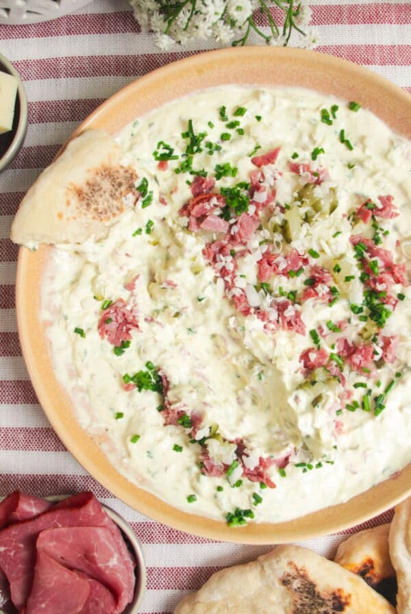 Creamy pickle dip in a pink bowl, with a piece of bread on the side.