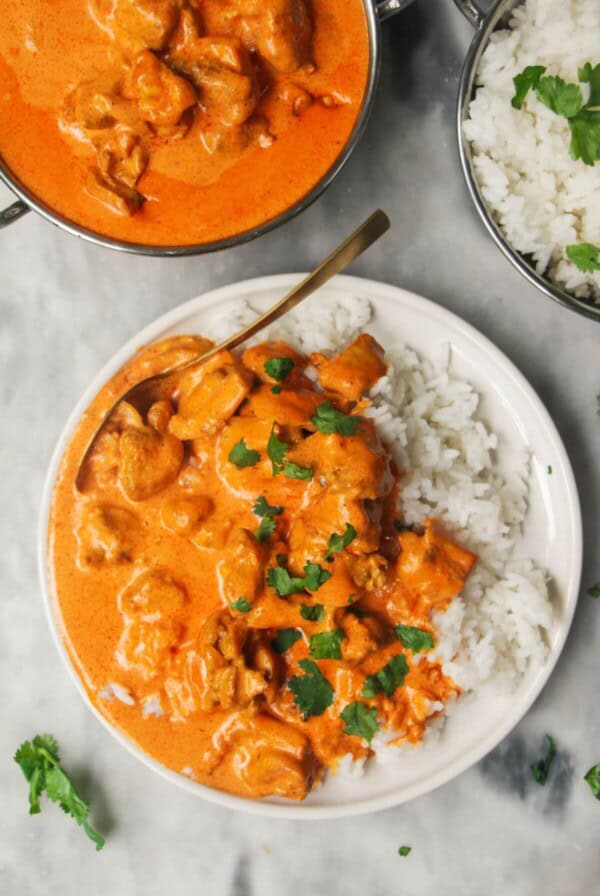 White plate with rice and butter chicken with bowl of butter chicken in the background and garlic naan.