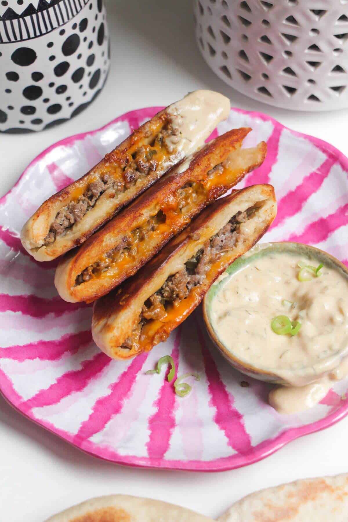 Three cheeseburger flatbreads on a pink and whiteplate with burger sauce on the side.